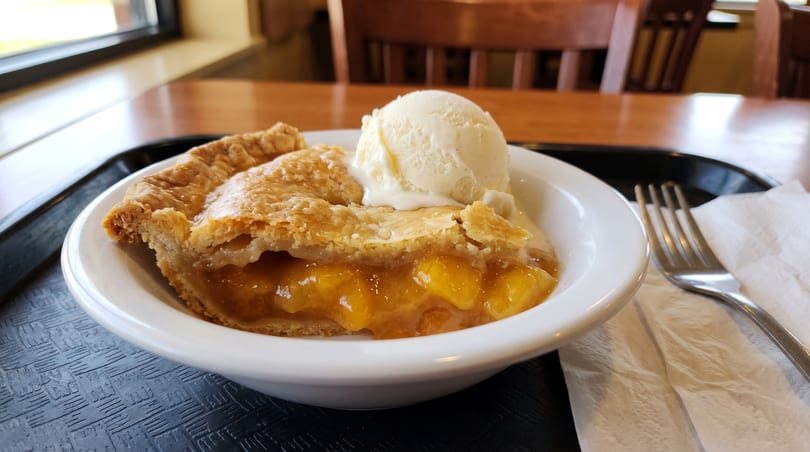 A slice of peach pie with a scoop of vanilla ice cream in a white bowl on a black tray.