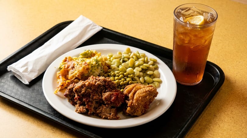 A cafeteria tray with meatloaf, fried chicken, lima beans, broccoli casserole, and iced tea.