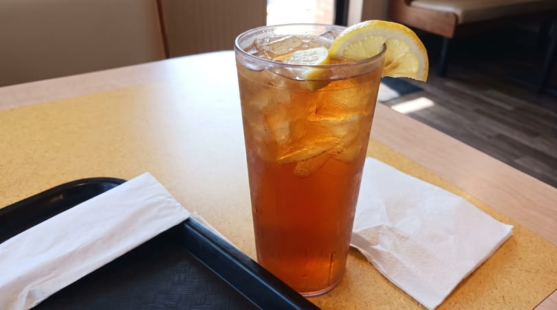 A tall glass of iced tea with a lemon slice sits on a restaurant table next to a black tray.