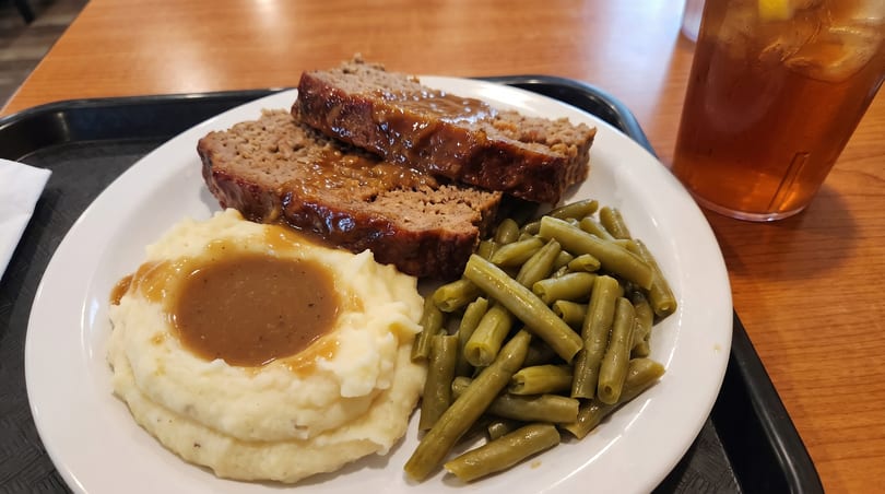 Meatloaf with gravy, mashed potatoes, and green beans on a white plate with iced tea.
